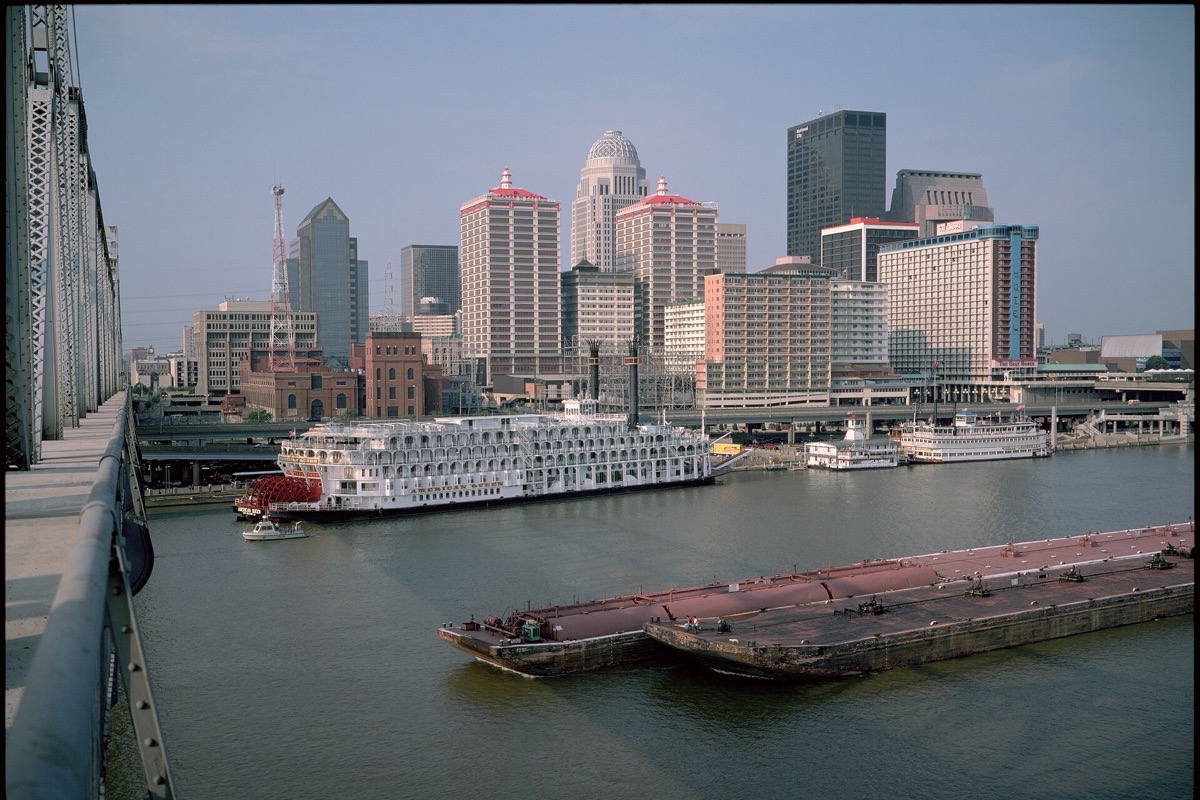 A towboat pushing a loaded barge past the Louisville waterfront on the Ohio, a white excursion boat moored at the wharf and the city skyline behind in soft haze.