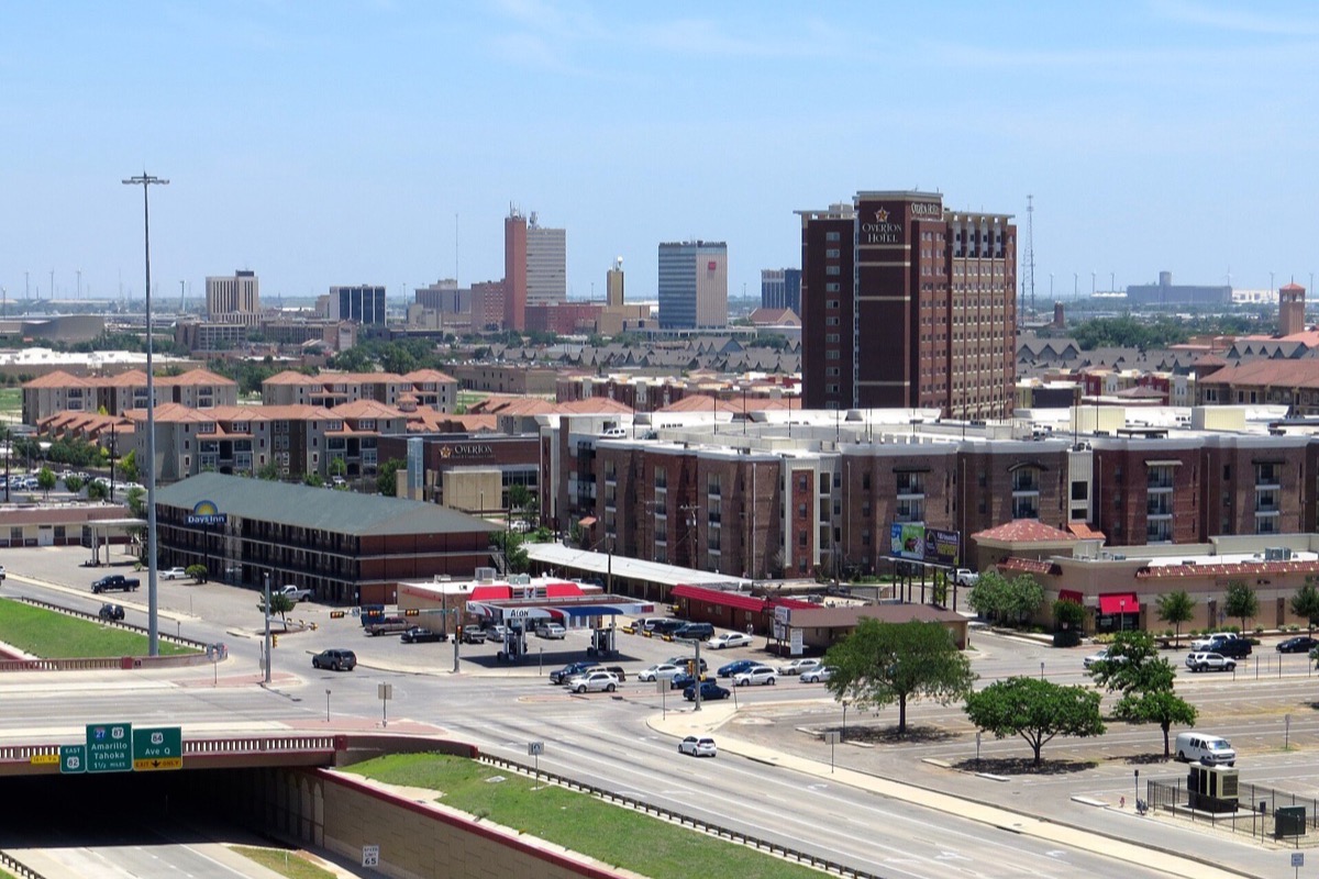 The Lubbock skyline under a clear summer sky, low brick apartment blocks and a scatter of office towers standing above a sunken road running through the middle of the frame.