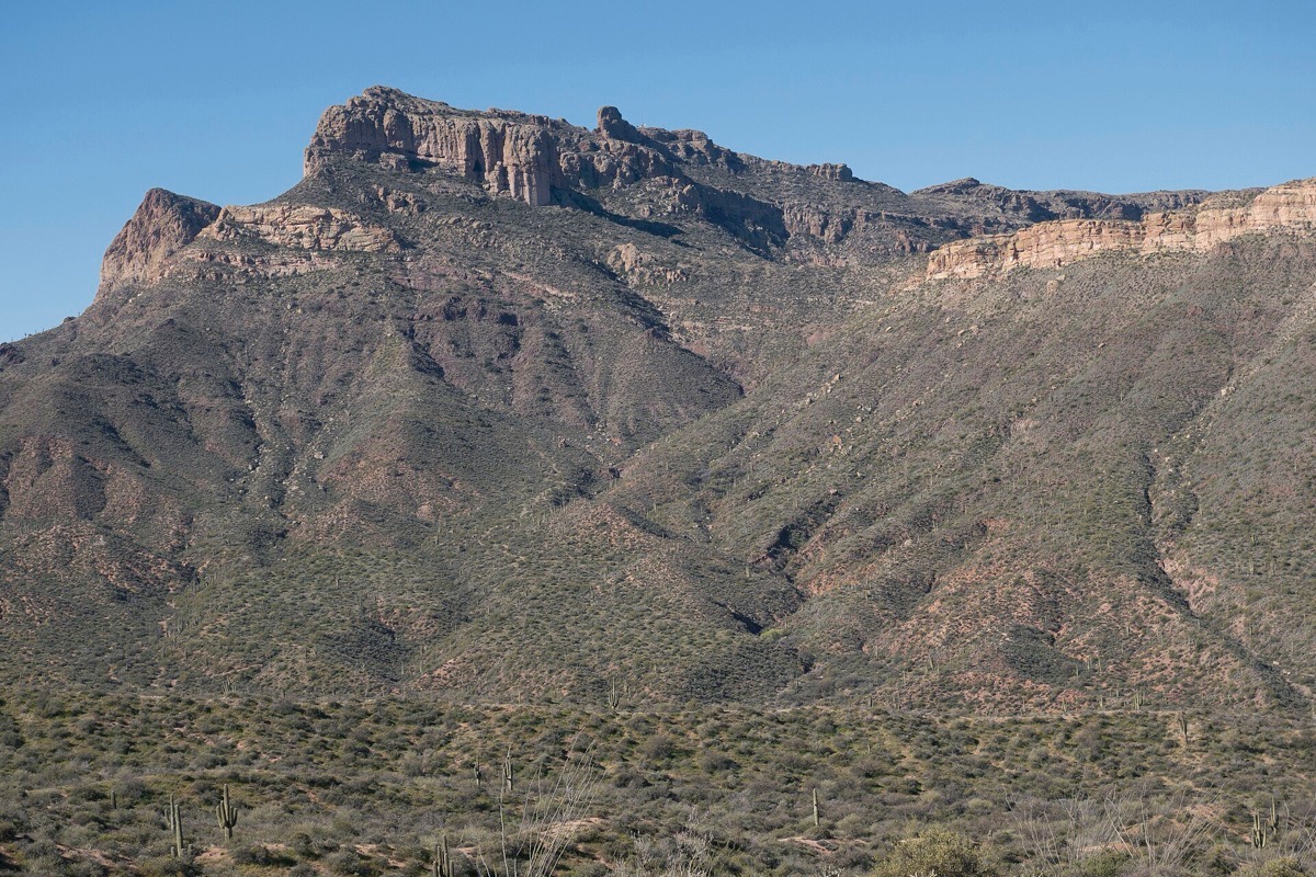 A long wall of Arizona desert mountain rising above Mesa in bands of grey and brown rock, the lower slopes covered in scrub and saguaro under a clear sky.