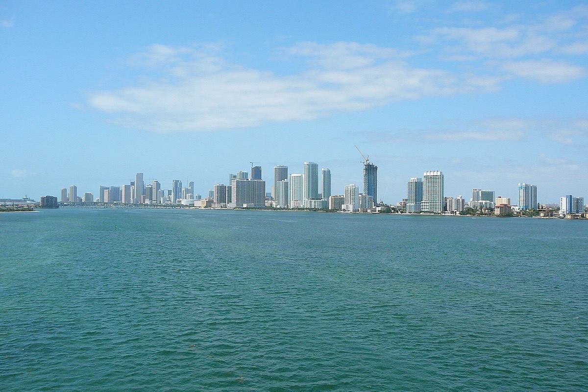 The Miami skyline seen from far out on Biscayne Bay, the towers reduced to a pale line on the horizon and bright green water filling most of the frame.