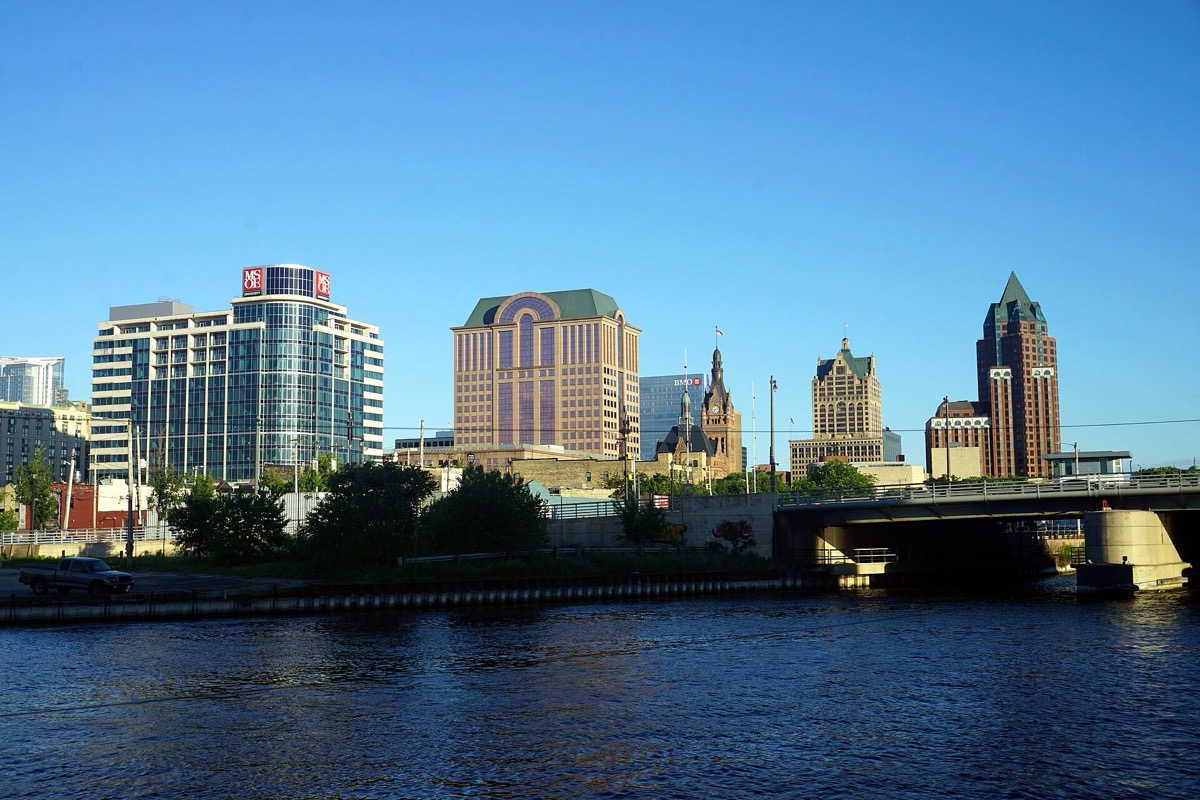 The Milwaukee river with a road bridge across it, brick and stone buildings and the city towers standing along the far bank, tall white cloud above them.