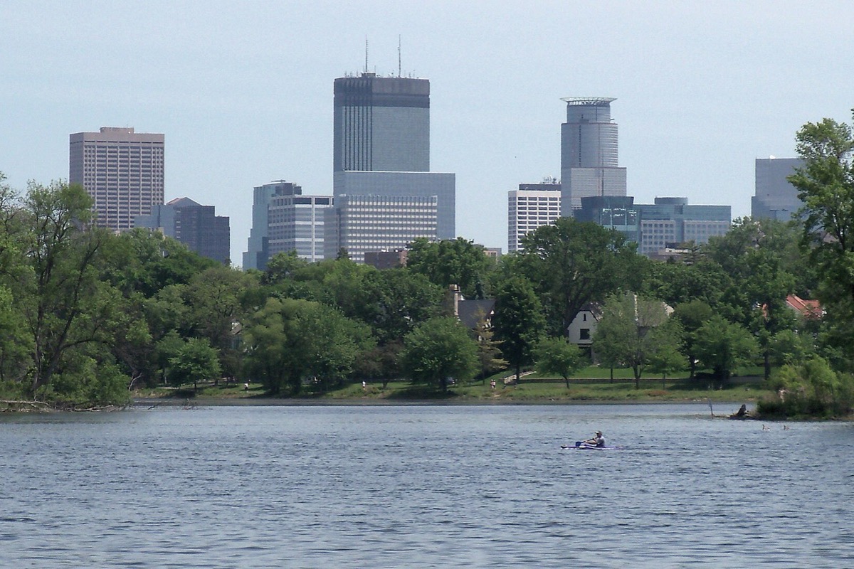 The Minneapolis skyline seen across one of the city lakes, a wooded shoreline with houses between, and a single rowing boat out on the flat water.