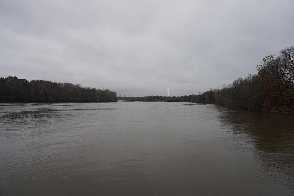 The Alabama River at Montgomery, wide and slow under a flat grey winter sky, with bare wooded banks closing in on both sides and a low road bridge and a tall chimney visible at the far bend.
