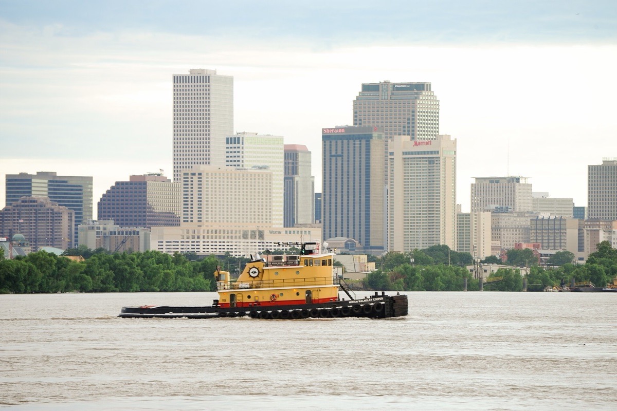 A yellow towboat working down the Mississippi past New Orleans, the brown river filling the foreground and the office towers of the city standing along the far bank in pale haze.