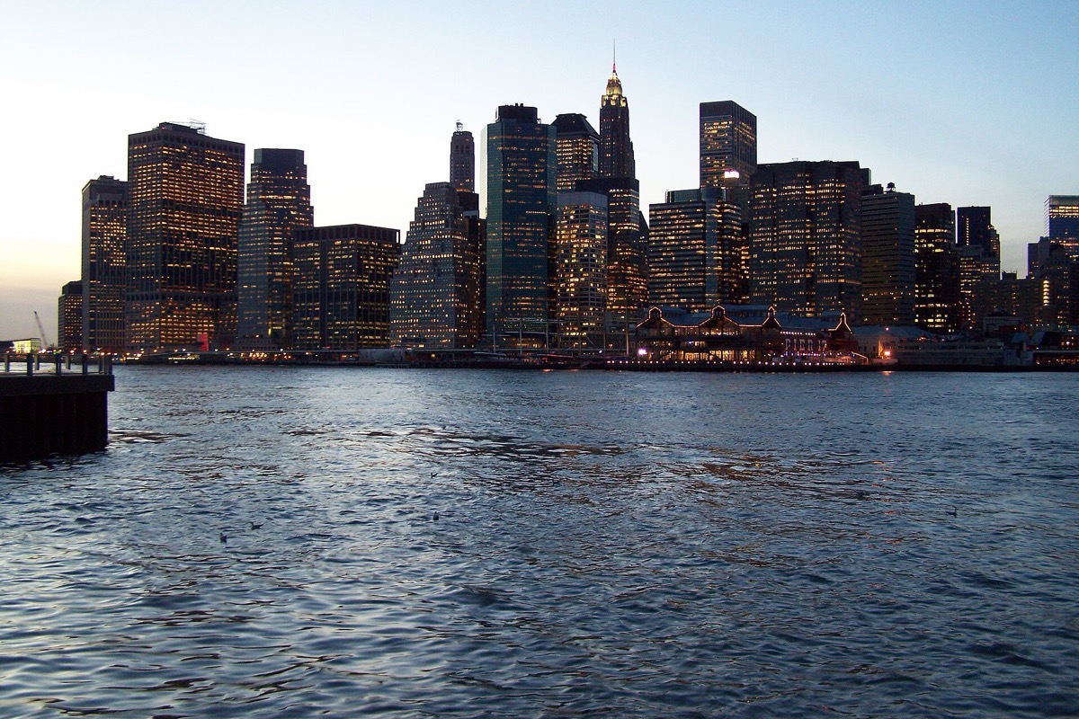 Lower Manhattan at dusk seen from the East River, the towers packed tight and lit from within, the water dark and choppy in front and the last daylight still in the sky.