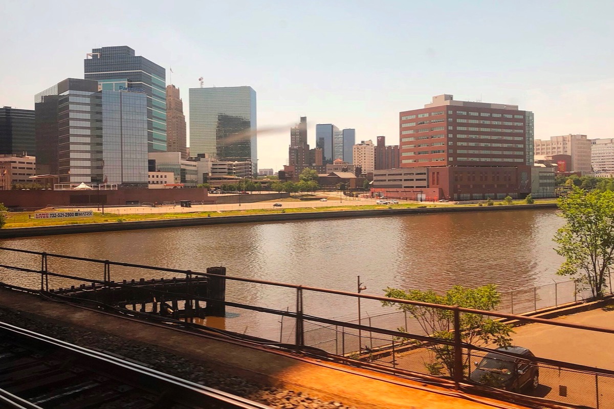 The Newark skyline seen from the rail line along the Passaic, office blocks and a tall brick building on the far bank, the river lit gold by low sun and the tracks running away in front.