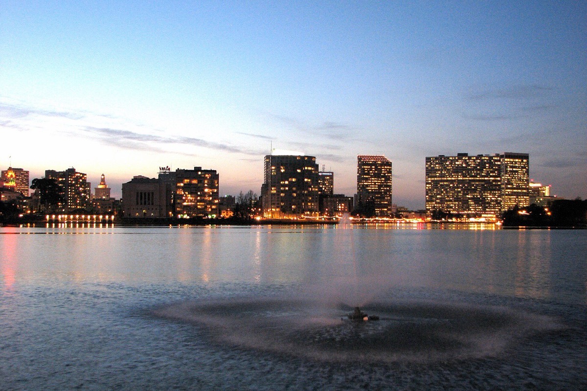 Lake Merritt in Oakland at dusk, the fountain working in the middle of flat water, the ring of lights around the shore and the office towers behind them under a pink and blue sky.