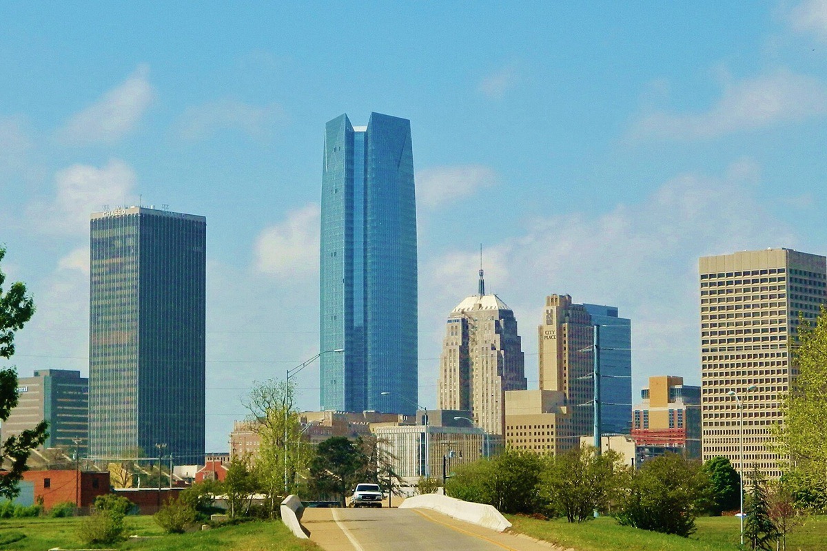 The Oklahoma City skyline from a road running into it, the tall glass tower in the centre and lower blocks around it, with new trees and green verges in the foreground under a clear sky.
