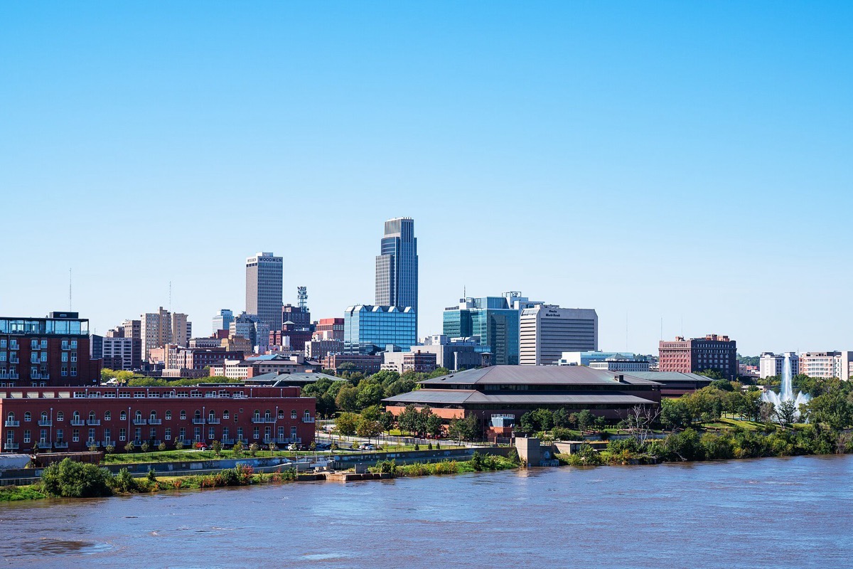 The Omaha skyline along the Missouri, brick warehouses and newer towers standing above the riverbank, with the wide brown river filling the foreground.