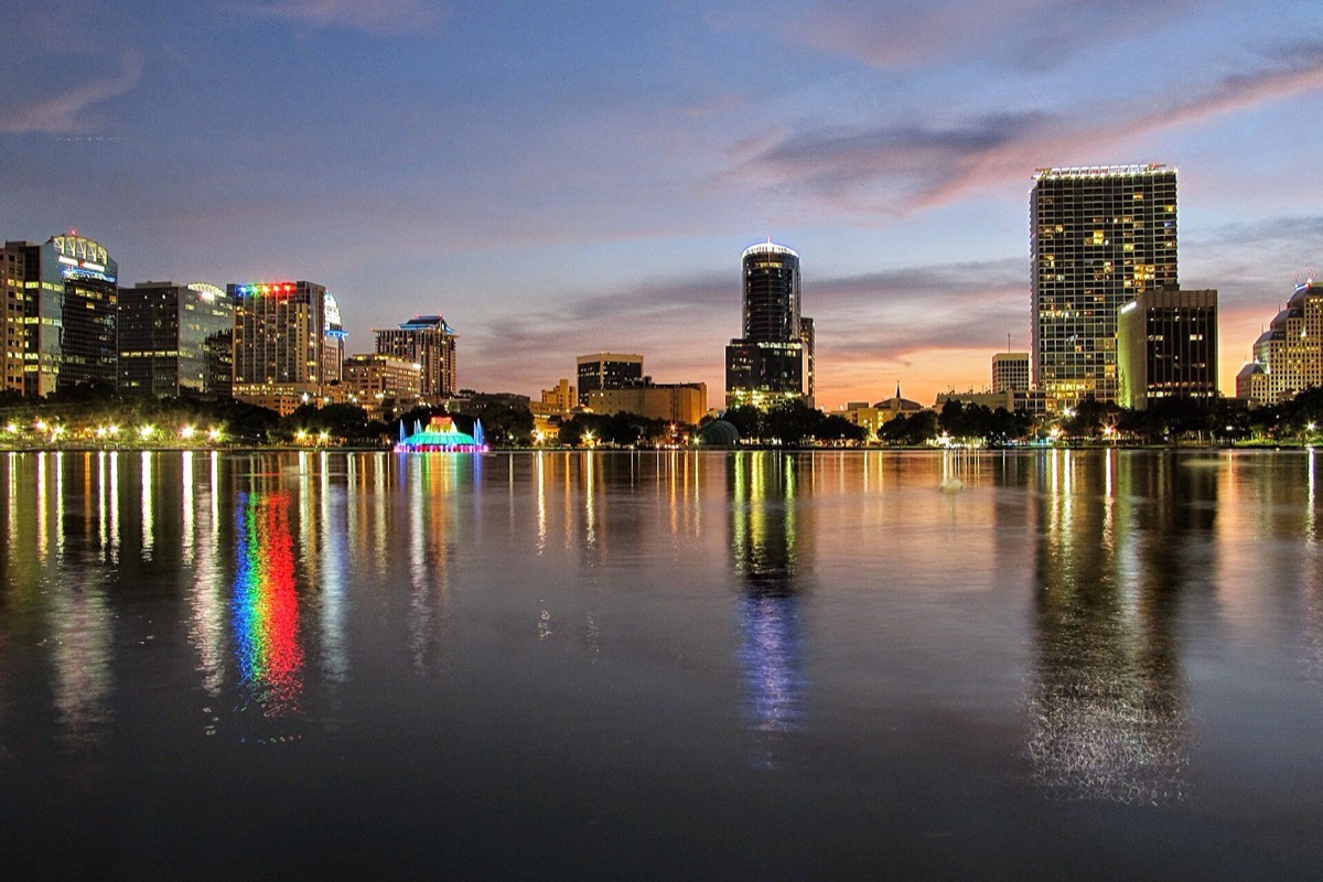 Lake Eola in Orlando at dusk, the lit fountain in the middle of the water throwing a band of colour across it and the city towers standing along the far shore under a pink sky.