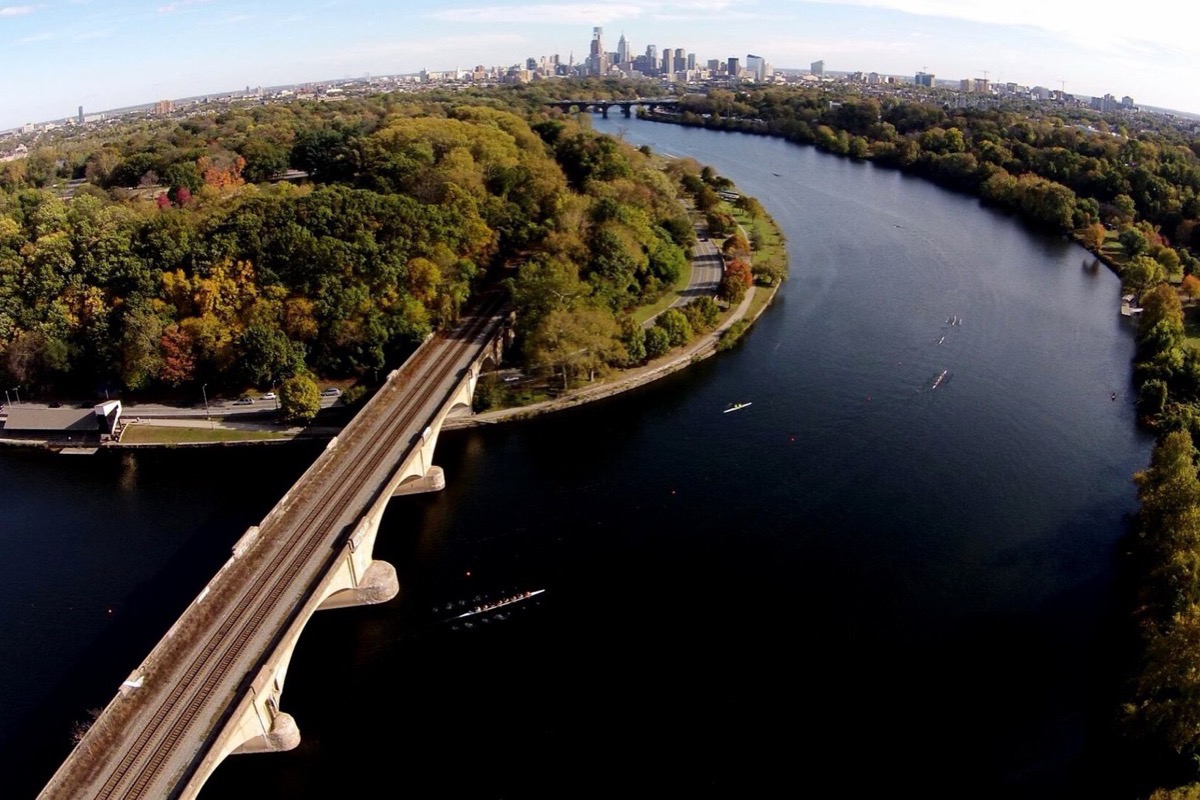 A bend of the Schuylkill in Philadelphia seen from above in autumn, a stone bridge crossing the near end, wooded parkland turning yellow inside the curve and the city skyline standing on the horizon.