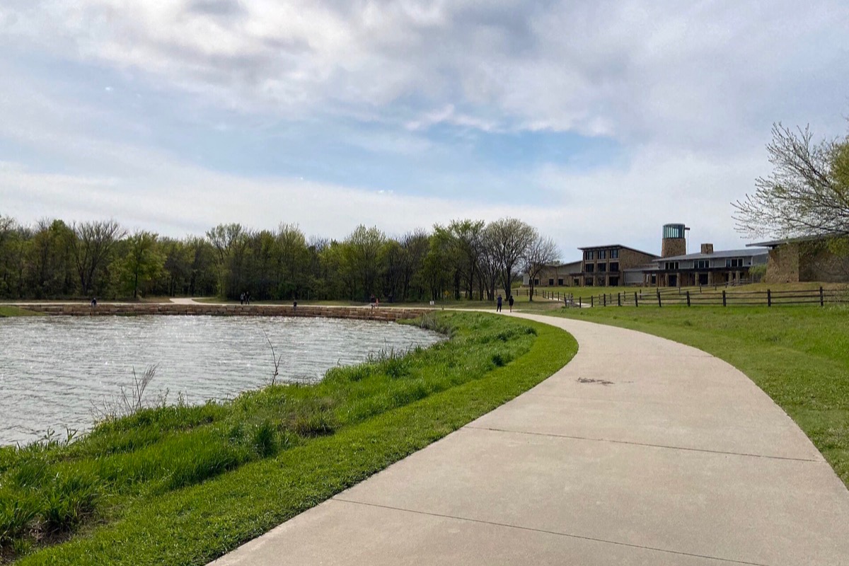 A paved trail curving along the edge of a lake in Plano, mown grass on both sides, a bare treeline along the far shore and a low timber and stone building among the trees.