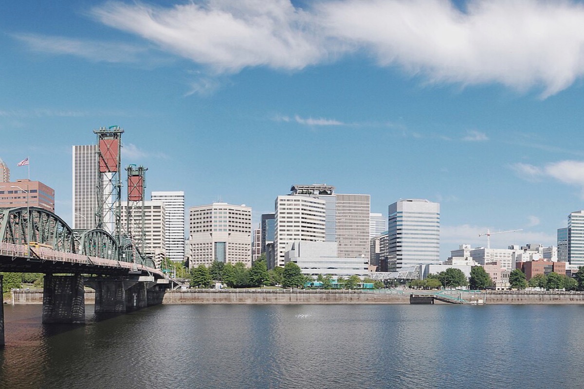 The Portland skyline across the Willamette with the steel bridge crossing the river in front of it, the water flat and a bank of white cloud above the buildings.