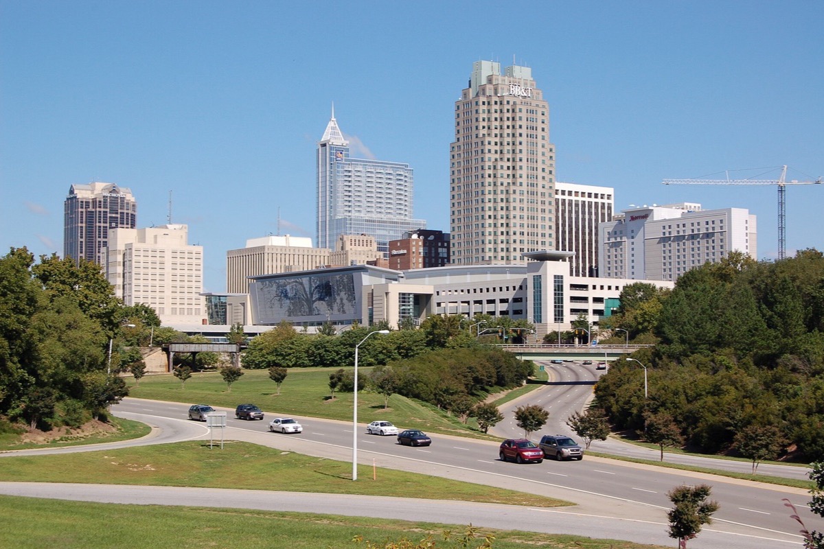 The Raleigh skyline seen from a rise across parkland and a road, stone and glass towers grouped close together with mature trees along the near side.