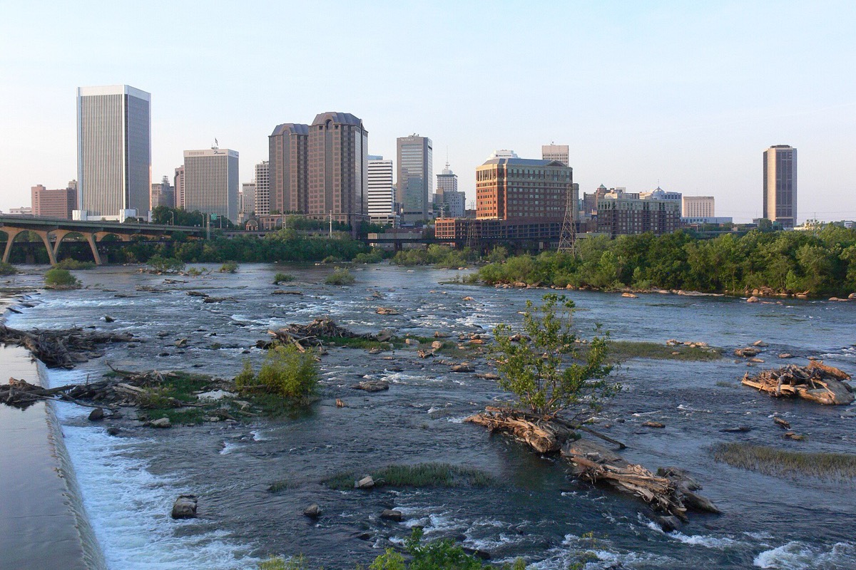 The James River running in shallow rapids over rock and around small wooded islets, with the Richmond skyline and a low road bridge along the far bank under a pale sky.