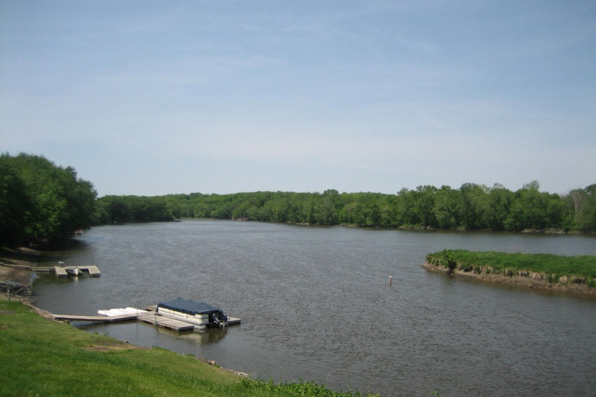 The Rock River in summer, a small covered pontoon boat moored at a floating dock on the near bank, dense green woodland along both shores and a grass bank in the foreground.