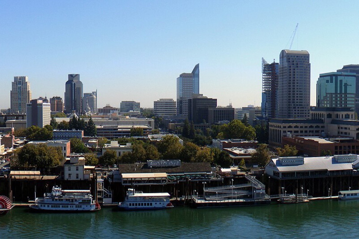 The Sacramento waterfront with two moored riverboats along the near bank, the office towers of the city behind them and a construction crane working among them.