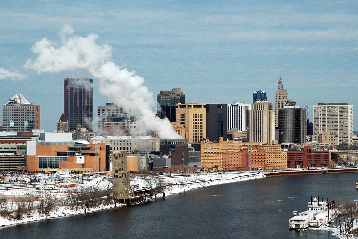 The Saint Paul skyline in deep winter, snow along both banks of the Mississippi, steam standing straight up from a heating plant and the office towers grouped behind it under a blue sky.