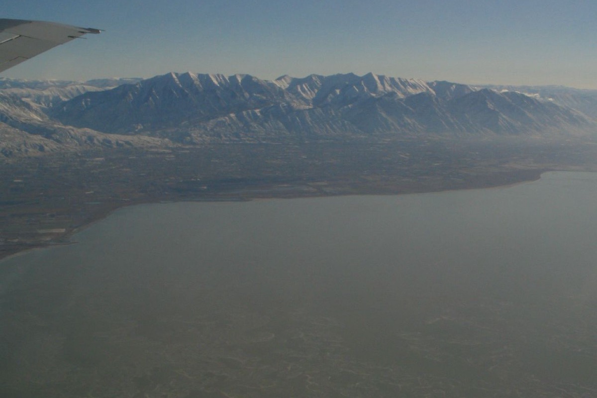 An aerial view along the Wasatch Front, the flat developed valley floor running between a wide lake in the foreground and a long snow topped range behind, with an aircraft wing at the top of the frame.