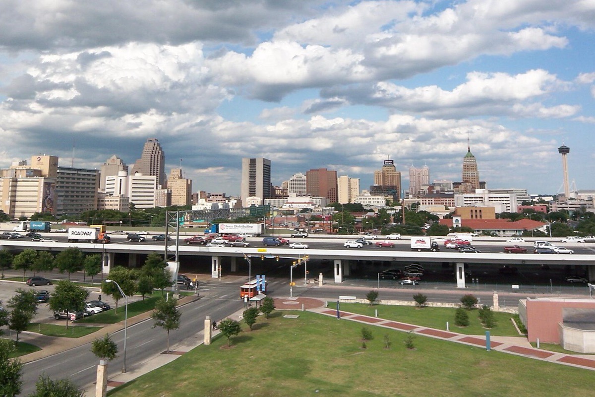 The San Antonio skyline across a green open space and a road, the tower with the observation top standing at the right hand end, under a sky full of broken white cloud.