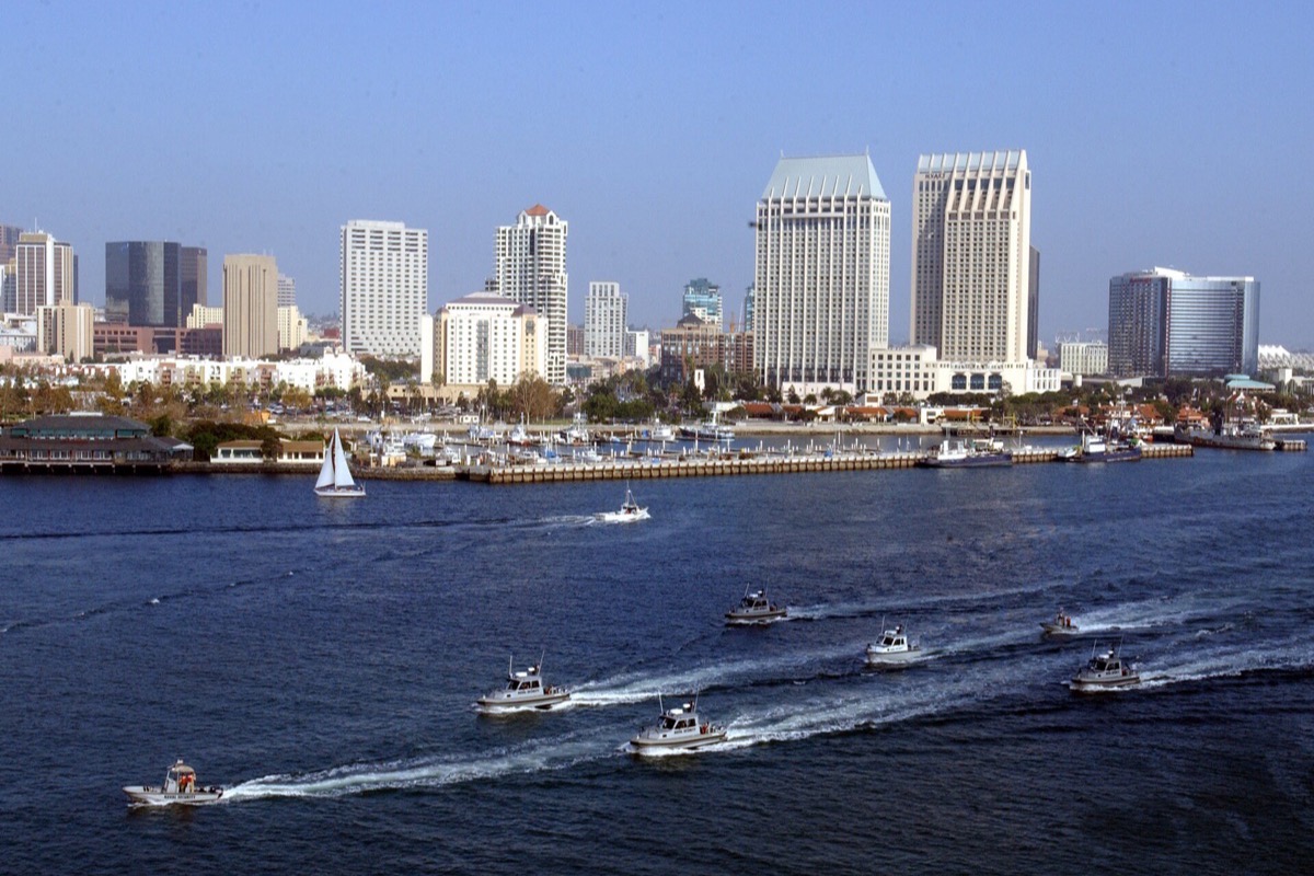 The San Diego bayfront seen from the water, hotel and office towers along the shore with masts of a marina in front of them, and small boats running across the bay leaving wakes.