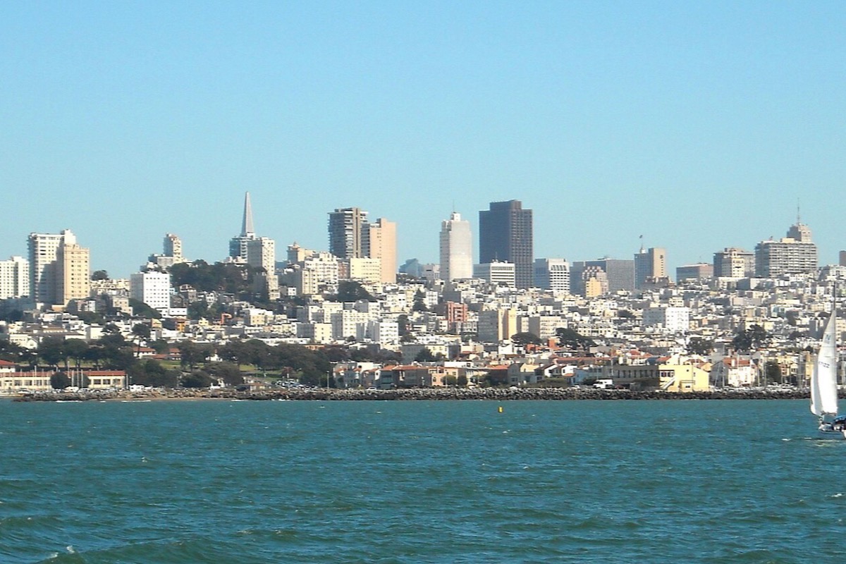 San Francisco seen low across the bay, the white city climbing its hills with the pyramid tower among the taller blocks, and flat blue green water filling the foreground.