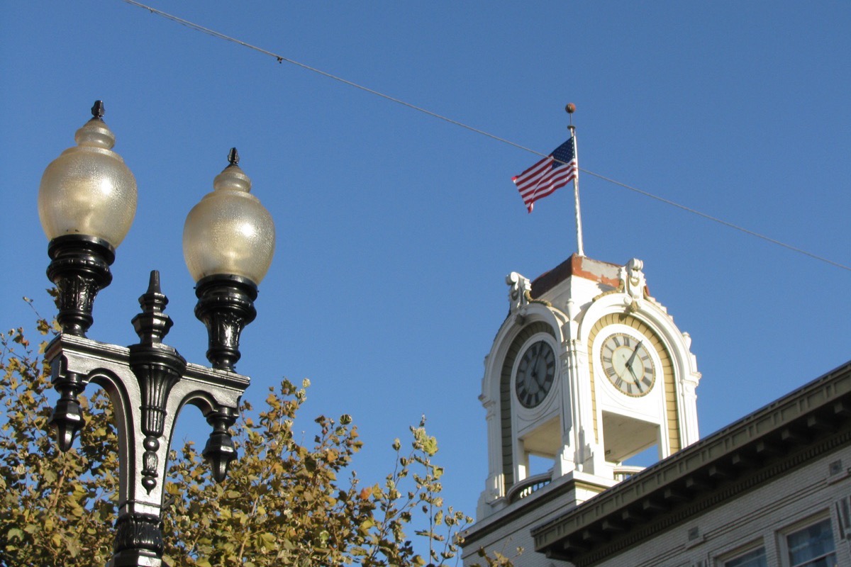A white clock tower with a flag on a historic Santa Ana commercial building, seen from the pavement below past an ornate twin lamp standard, against a clear blue sky.