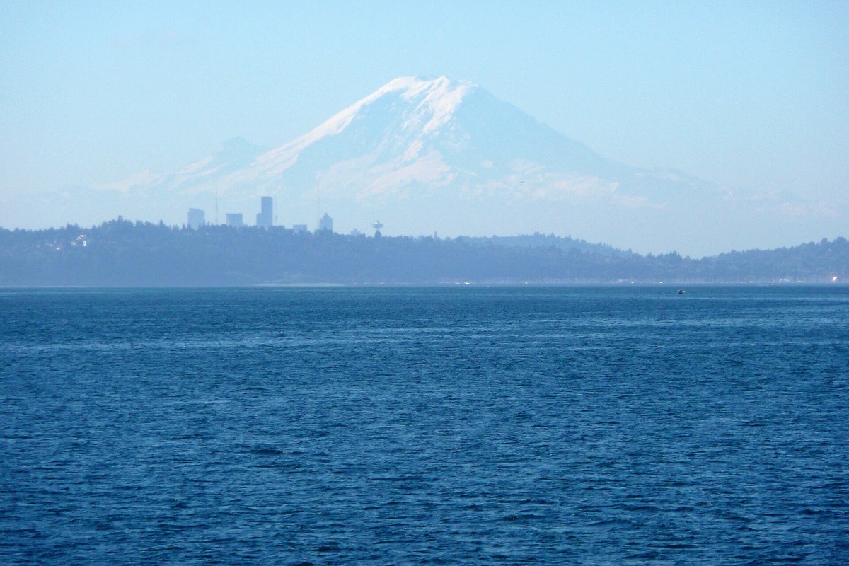 Mount Rainier standing white and enormous on the horizon beyond Seattle, the city itself reduced to a thin pale line on the water, with deep blue sound filling the foreground.