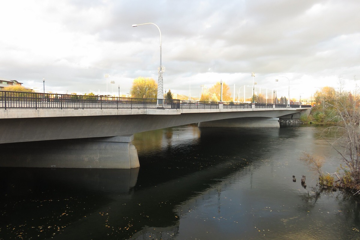 A concrete road bridge crossing the Spokane River, seen low from the bank, with autumn trees and floodlight towers beyond and fallen leaves scattered across dark water.