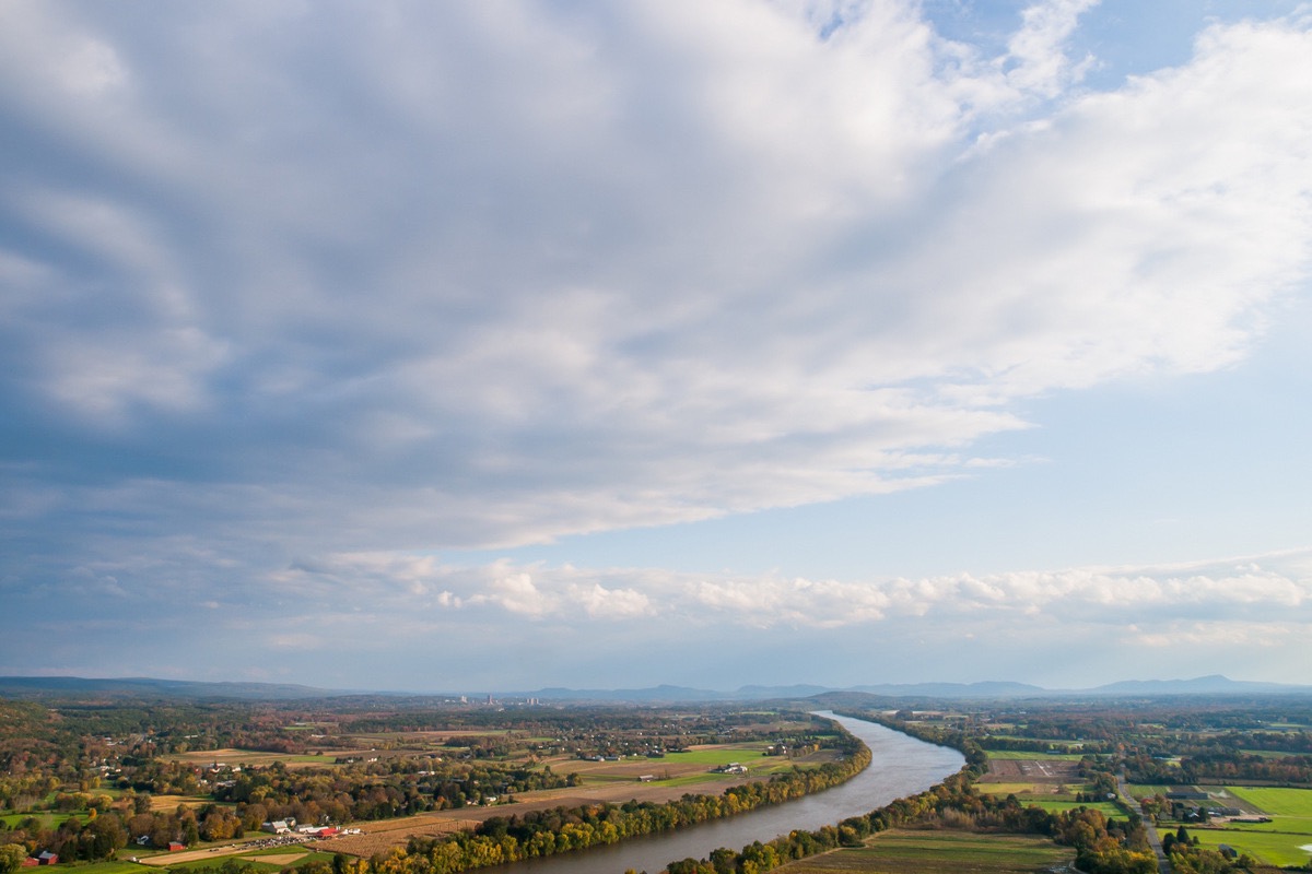 The Connecticut River seen from high ground, winding between farmland and scattered settlement under a wide sky of broken cloud, with low hills along the horizon.