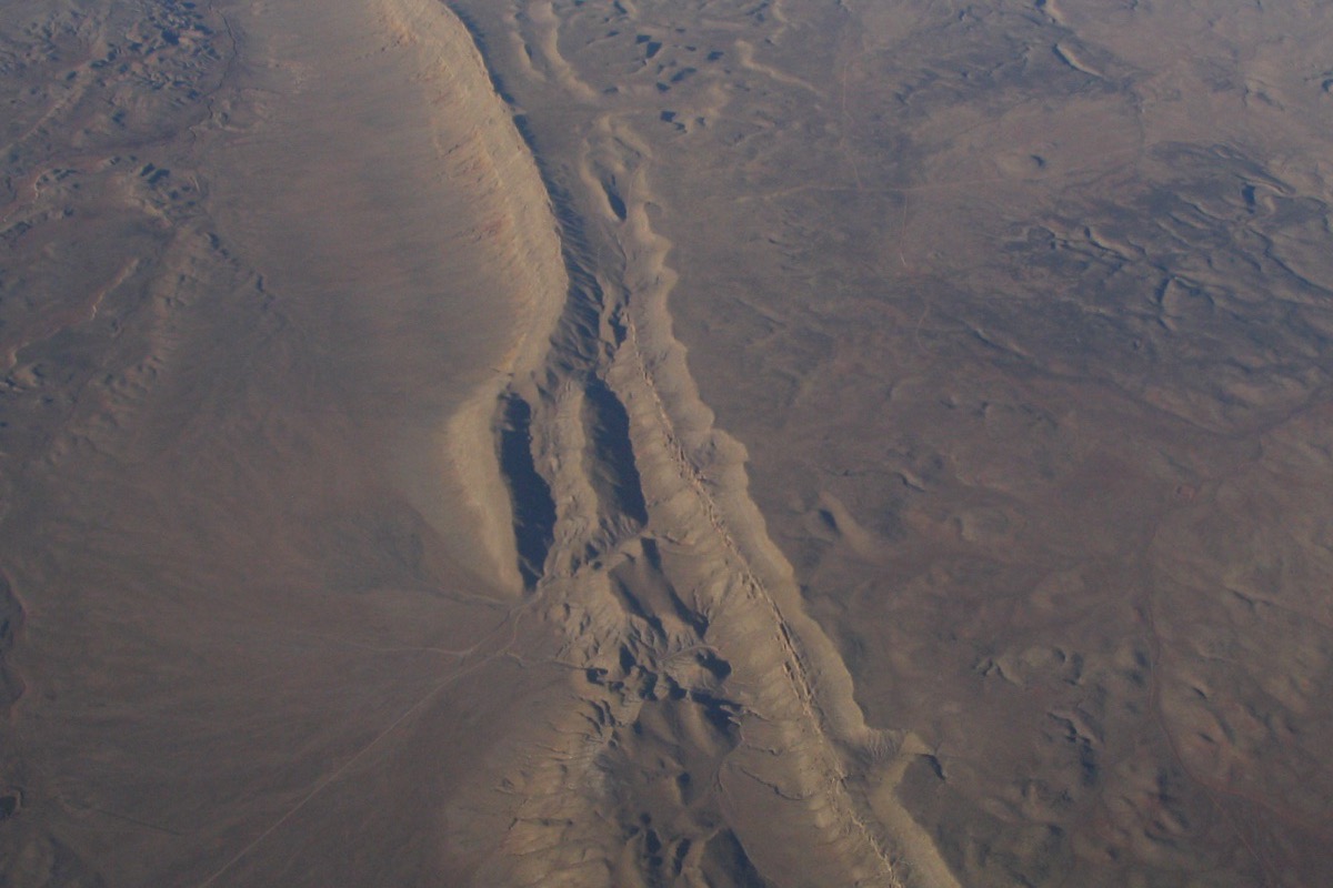An aerial view of a long fault escarpment cutting diagonally across empty high desert northeast of St. George, with eroded gullies along its face and faint tracks running out across the flat ground on either side.