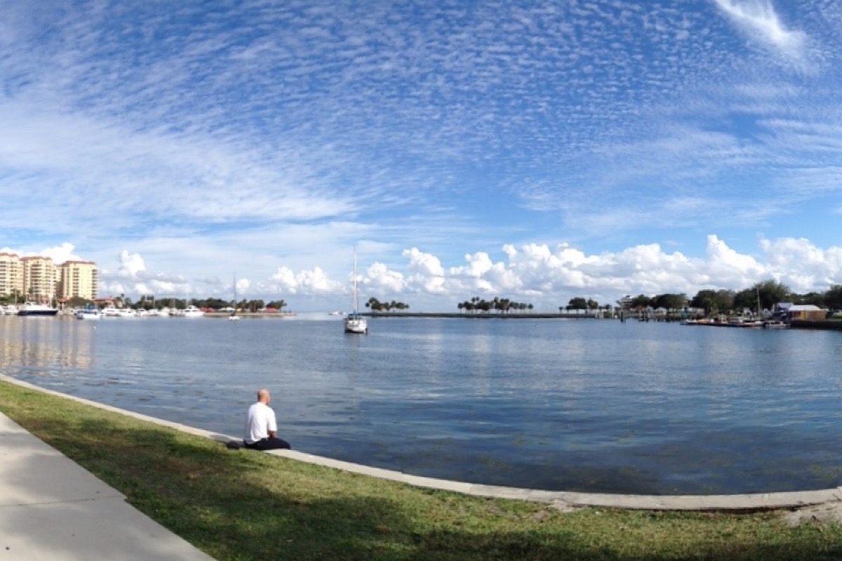 The St. Petersburg waterfront on a bright day, moored yachts and a marina along the left shore, a low green spit running across the middle distance and calm bay water under a wide mackerel sky.