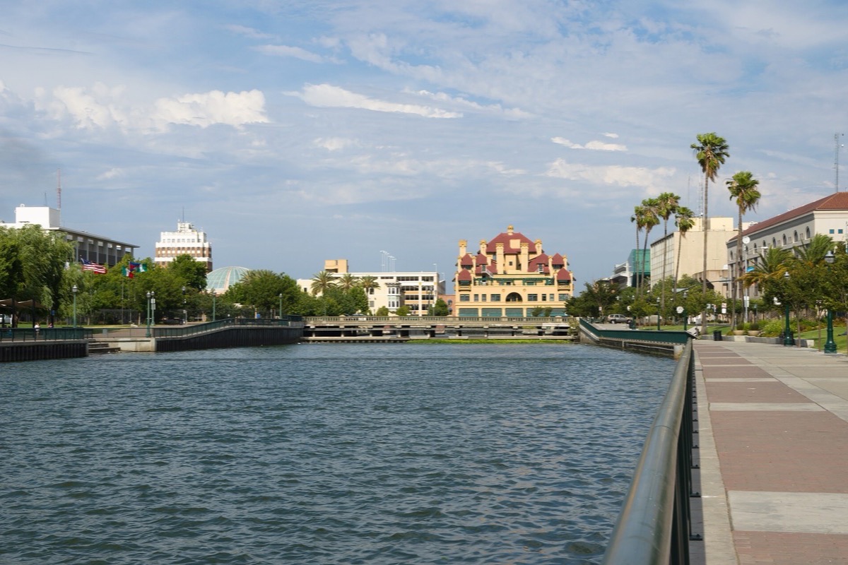 The Stockton waterfront channel, flat water between concrete walls with a paved walkway and palms along the near side and the ornate roof of an older building on the far bank.