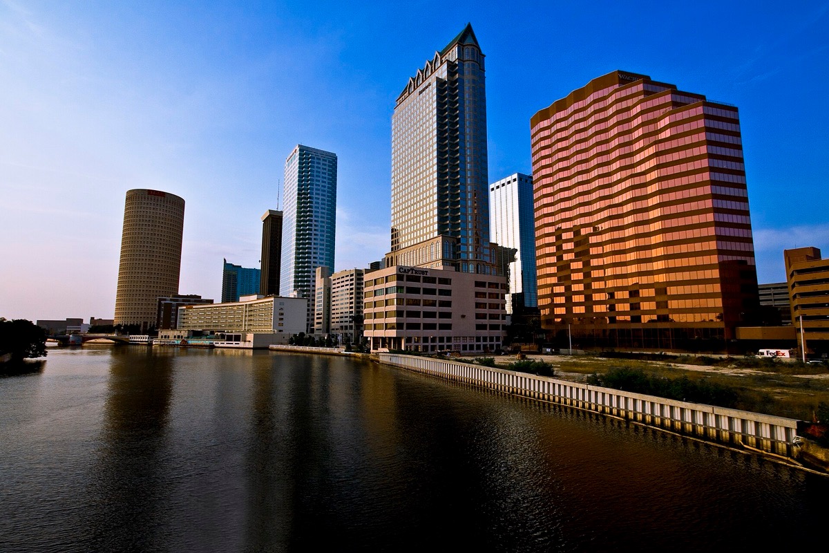 The Tampa skyline at dusk seen from across the river, a cylindrical tower and a bronze glass block among the others, all of it reflected in dark still water.