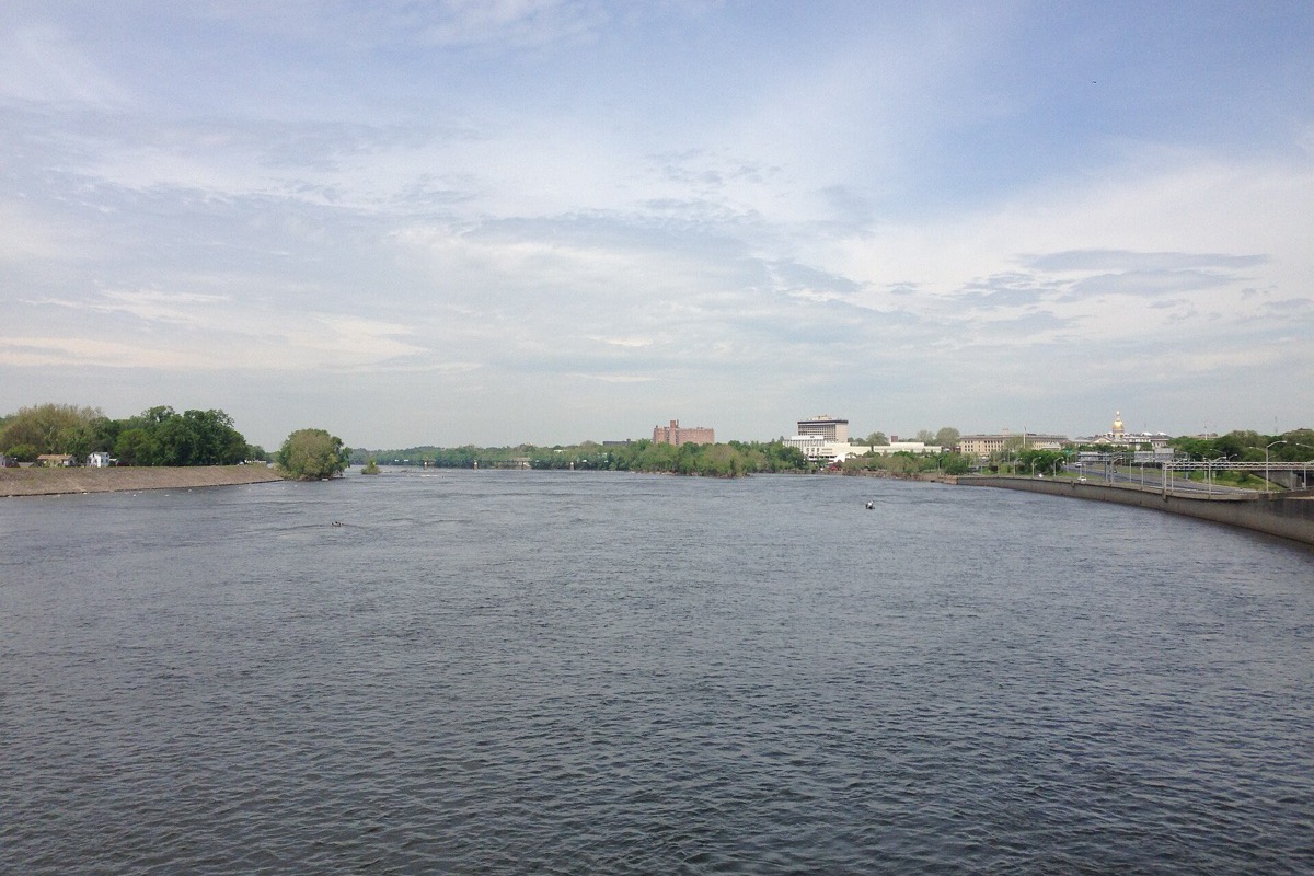 The Delaware River seen from a bridge at Trenton, wide grey water running to a low treeline, with the capital's buildings and a gold dome along the right bank under a hazy sky.