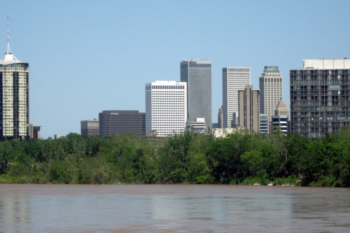 The Tulsa skyline seen from across the Arkansas, a broad brown river in the foreground, a wooded bank between, and the office towers standing above the trees.