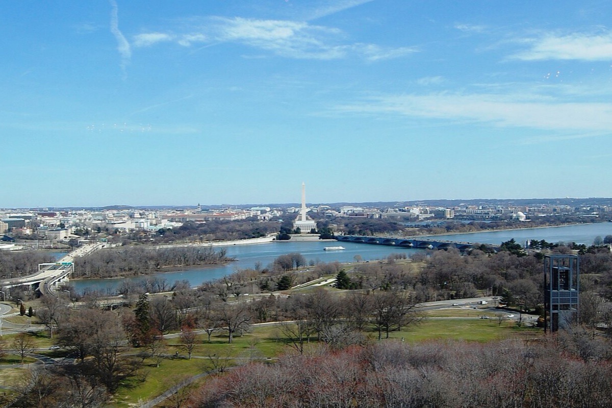 Washington seen from the high ground across the Potomac in winter, the river and its bridges in the middle, the monument and the long green of the Mall beyond, and bare trees in the foreground.