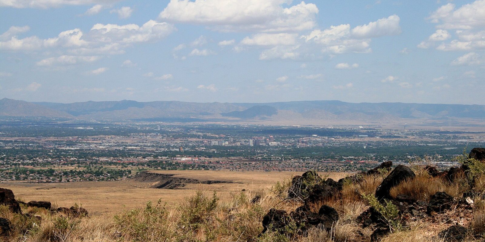Albuquerque's low downtown cluster seen across a dry grassy valley from a rocky foothill, with a long mountain ridge and scattered clouds behind