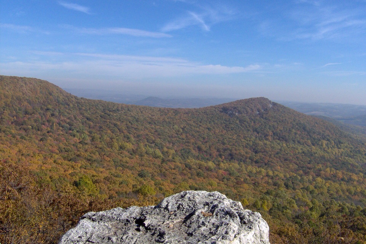 A long forested Pennsylvania ridge running away into autumn haze, seen past a slab of pale rock on the summit under a clear blue sky.