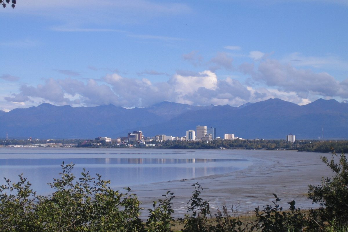 Anchorage's low downtown skyline sits across the tidal mudflats of Knik Arm, with the Chugach Mountains rising behind under scattered clouds.