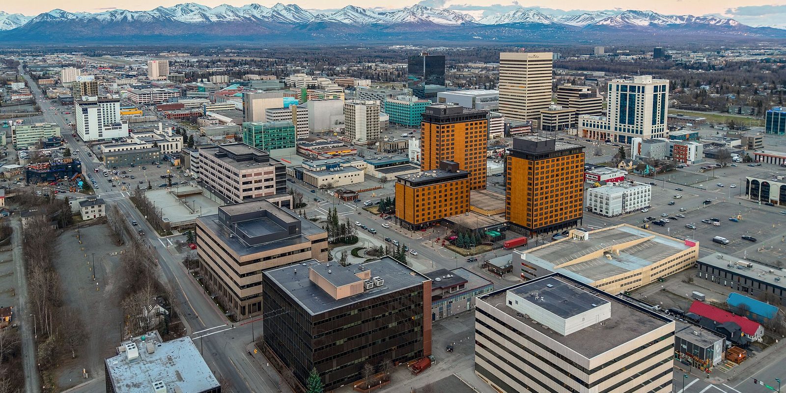 Aerial over downtown Anchorage's mid-rise blocks and two amber-clad towers, with bare trees and a long snow-capped mountain range across the horizon