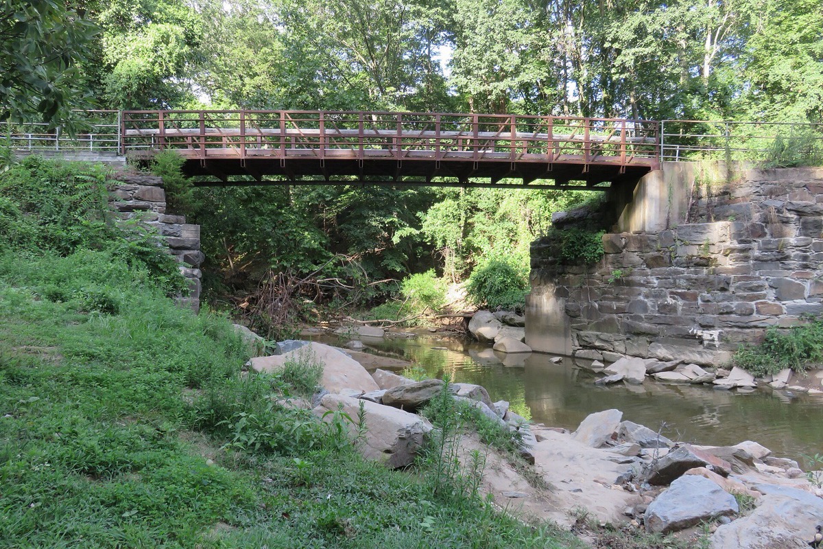 A low timber trail bridge on stone abutments crossing a shallow rocky creek in a Virginia park, dense green summer woodland closing in on both banks.