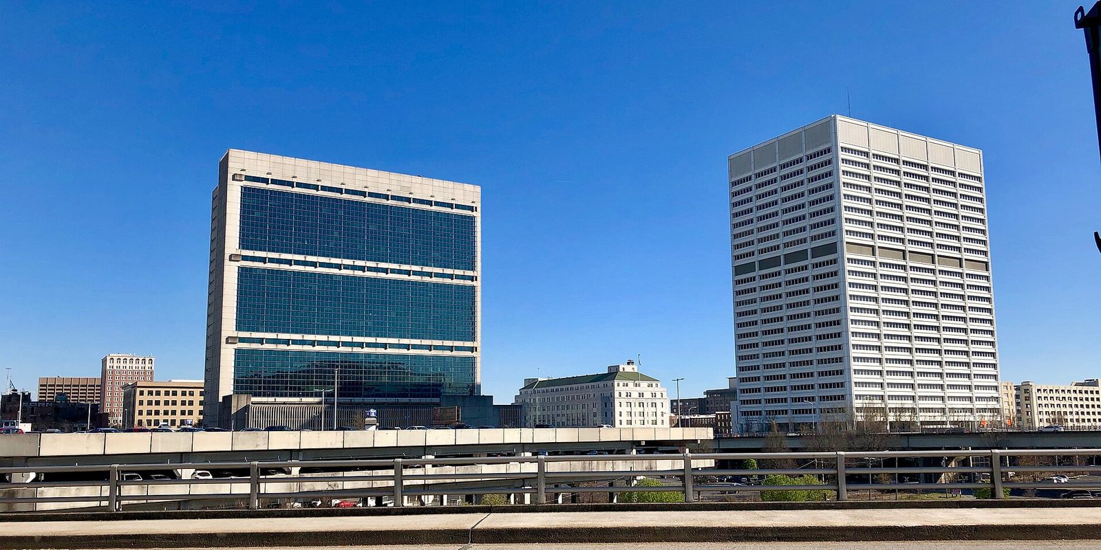 Two large Atlanta office towers rising beyond an elevated freeway and guardrails, seen from road level under a clear deep blue sky