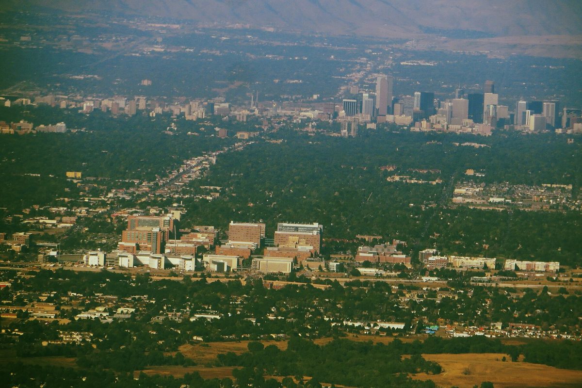 An aerial over Aurora shows a brick medical campus set among tree-filled neighborhoods, with Denver's skyline and the Front Range hazy beyond.