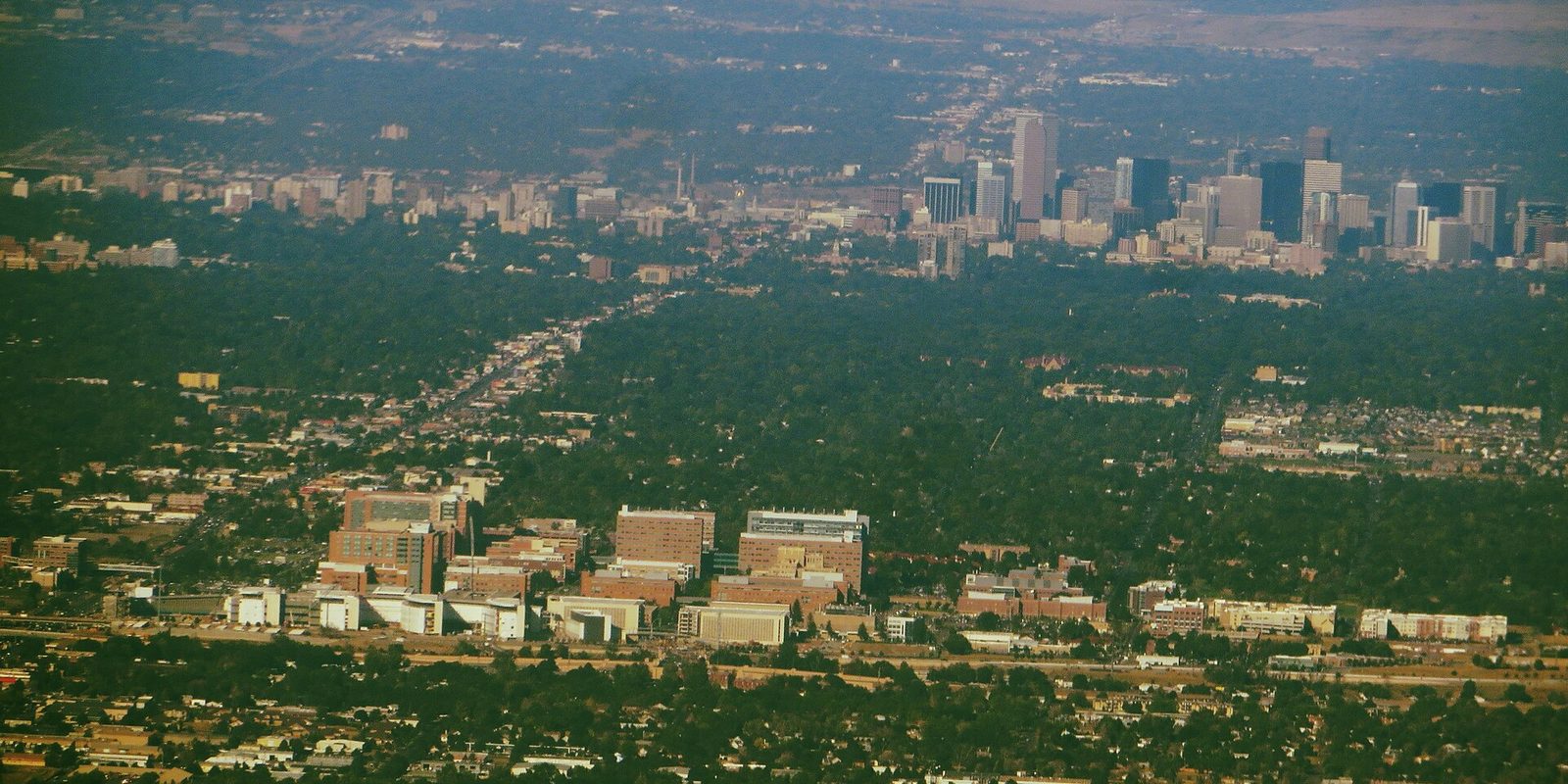 Hazy aerial across Aurora's tree-covered neighborhoods and a large brick medical campus, with Denver's skyline small on the far horizon