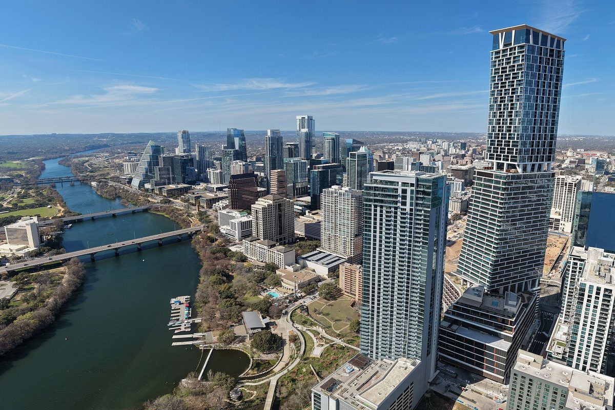 Downtown Austin towers rise above Lady Bird Lake, where bridges cross the water and bare trees line the banks under a clear blue sky.