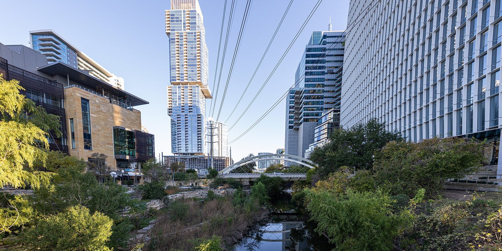 Austin's glass towers rising above a leafy downtown creek, with a white arched bridge crossing it and power lines overhead against a clear sky