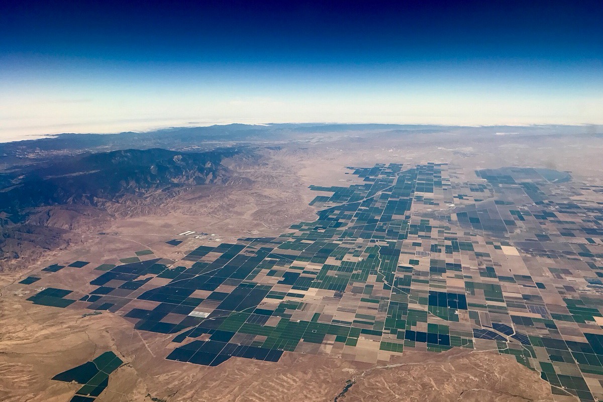 Kern County farmland from the air, a grid of green and bare fields running flat to a haze covered horizon with dry brown coastal hills along the near edge.