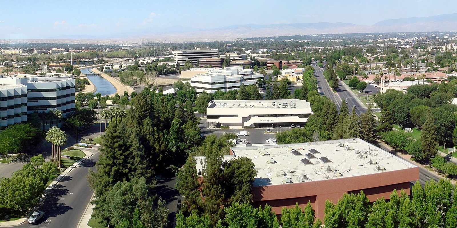 Bakersfield from above, low-rise office buildings and palms along a straight concrete canal and wide boulevard, with hazy hills on the horizon