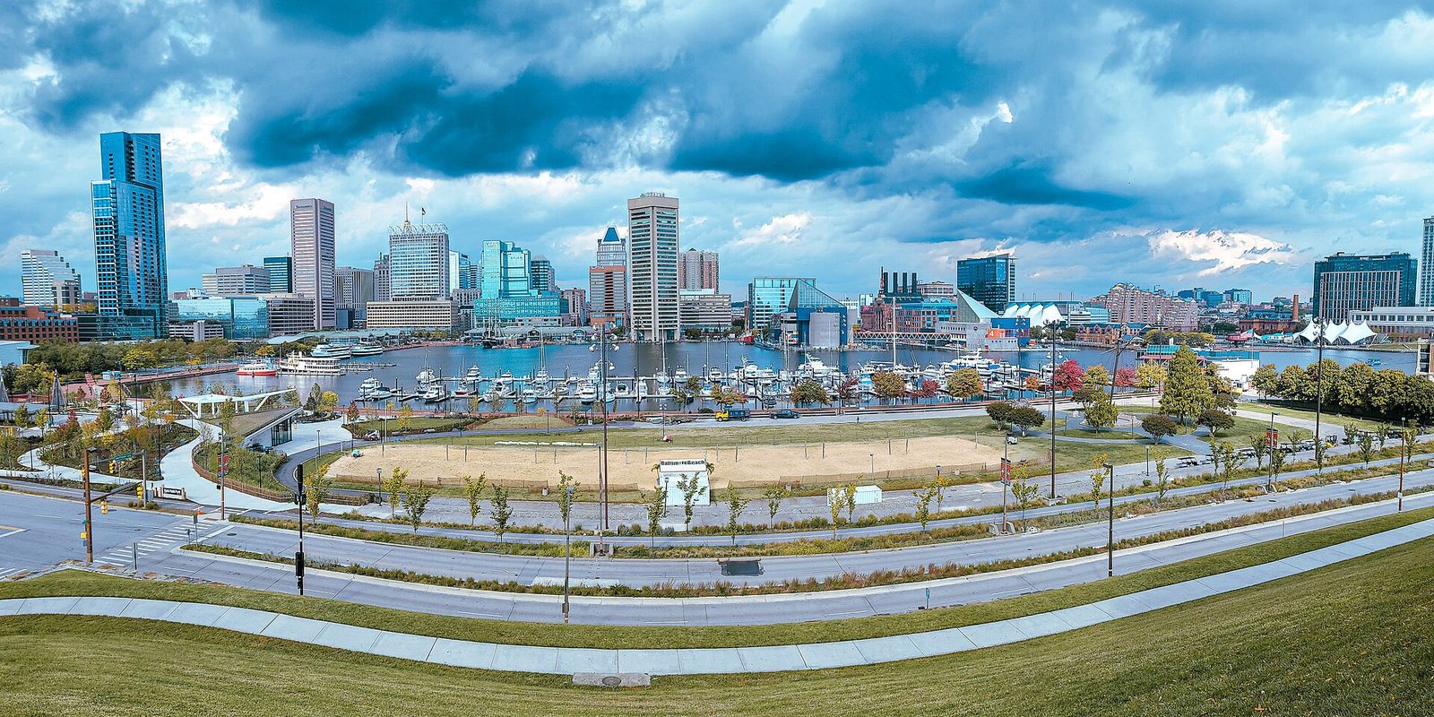 Baltimore's Inner Harbor viewed from a grassy rise, a marina packed with boats below the downtown skyline and heavy clouds breaking overhead